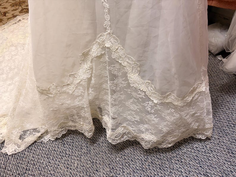 May include: Ivory-colored wedding dress with a lace hem. The dress features a layered design with a sheer overlay and intricate lace detailing along the bottom edge. The lace trim has a scalloped edge.