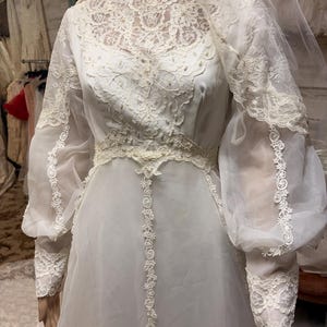 May include: A vintage white wedding dress with a high neckline and long, sheer sleeves. The bodice and sleeves feature intricate lace detailing, with a delicate floral pattern. The dress includes a matching veil.