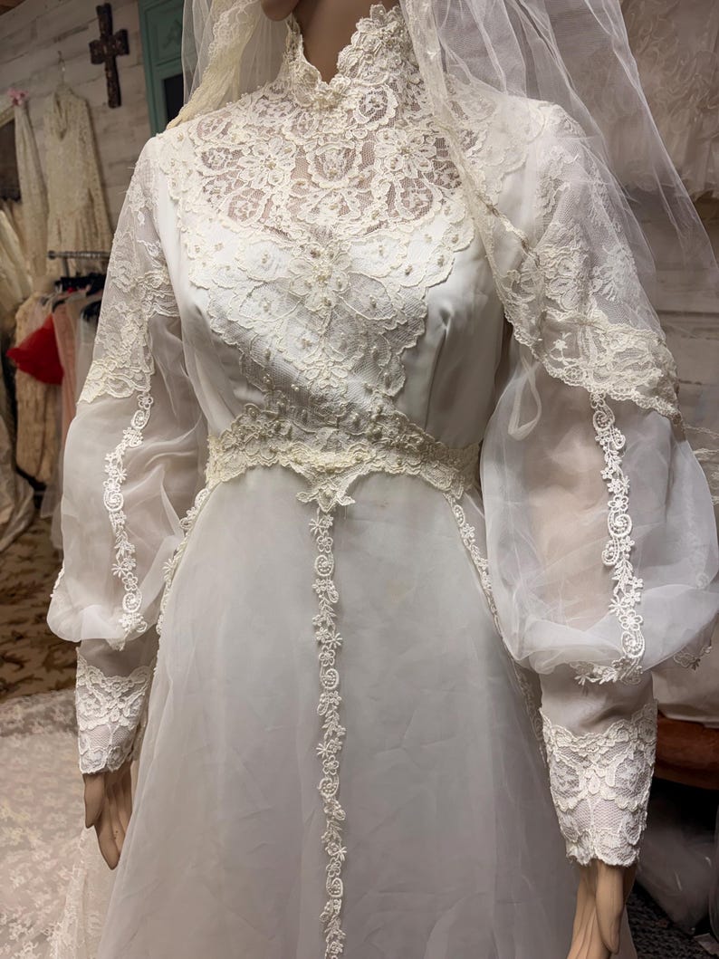 May include: A vintage white wedding dress with a high neckline and long, sheer sleeves. The bodice and sleeves feature intricate lace detailing, with a delicate floral pattern. The dress includes a matching veil.