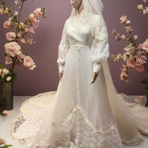 May include: A vintage-style wedding dress on a mannequin, featuring a high neckline, long sleeves, and a flowing skirt with a lace trim. The dress is white with delicate lace details. A long veil and floral headpiece complete the bridal look.