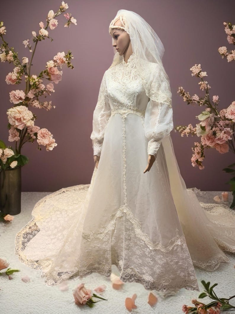 May include: A vintage-style wedding dress on a mannequin, featuring a high neckline, long sleeves, and a flowing skirt with a lace trim. The dress is white with delicate lace details. A long veil and floral headpiece complete the bridal look.