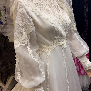 May include: Ivory vintage wedding dress with a high neckline and long, sheer sleeves. The bodice and sleeves feature intricate lace detailing. The dress has a flowing skirt and is paired with a matching veil. A classic bridal gown.