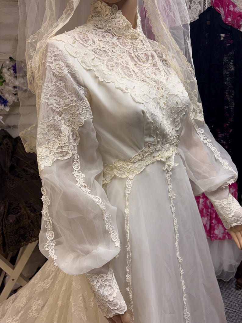 May include: Ivory vintage wedding dress with a high neckline and long, sheer sleeves. The bodice and sleeves feature intricate lace detailing. The dress has a flowing skirt and is paired with a matching veil. A classic bridal gown.