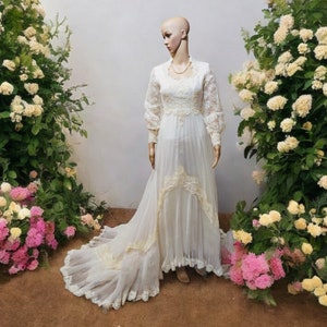 May include: A white lace wedding dress with long sleeves and a flowing skirt. The dress has a fitted bodice and a sweetheart neckline. The skirt is layered and has a scalloped hem.