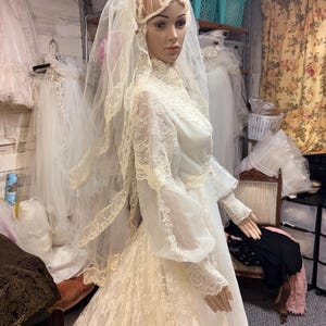May include: A vintage ivory wedding dress on a mannequin. The dress features long sleeves, a high neckline, and intricate lace detailing. A matching veil and headpiece complete the bridal ensemble. Other wedding dresses are visible in the background.