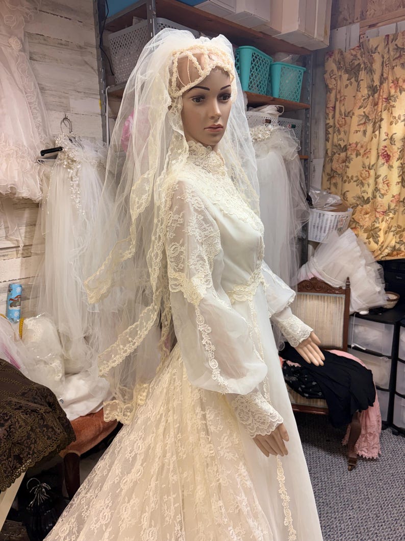 May include: A vintage ivory wedding dress on a mannequin. The dress features long sleeves, a high neckline, and intricate lace detailing. A matching veil and headpiece complete the bridal ensemble. Other wedding dresses are visible in the background.