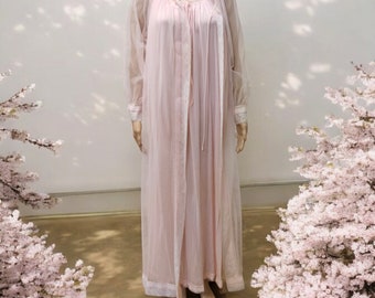 Op de afbeelding: Een lichtroze vintage nachtjapon en badjas set op een mannequin. De nachtjapon heeft een ronde halslijn met kanten rand, lange mouwen en een rok tot op de grond. De doorschijnende badjas heeft kanten rand en een striksluiting. Een parelketting wordt gedragen.