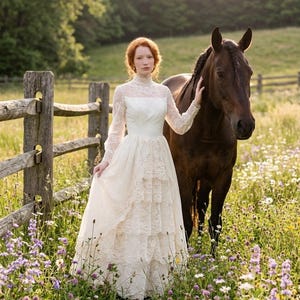 May include: A woman in a white lace wedding dress stands in a field of wildflowers next to a brown horse. The dress has long sleeves and a high neckline. The scene is set in a sunny meadow with a wooden fence in the background.