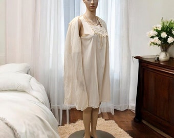 May include: A vintage ivory-colored nightgown and matching robe set displayed on a mannequin. The nightgown features lace detailing at the neckline and hem. A pearl necklace adds a touch of elegance. The scene is set in a bedroom with a bed and a wooden dresser.