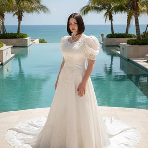 Puede incluir: Un vestido de novia blanco con una larga cola y detalles de encaje. El vestido presenta mangas cortas abullonadas y un cuello alto. La novia está de pie frente a una piscina con palmeras y el mar de fondo.