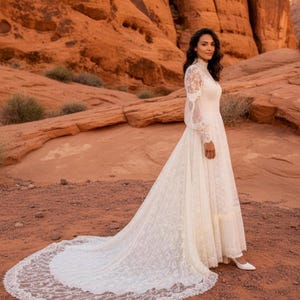 May include: A white lace wedding dress with a high neckline and long sleeves. The dress features a long train with a delicate lace trim. The bride is wearing white shoes. The background is a desert landscape with red rock formations.