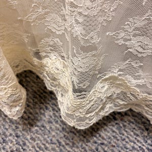 May include: Close-up of ivory lace fabric with a floral pattern. The delicate lace is draped, showing the intricate details and texture. The edges are scalloped, adding a decorative touch. The fabric is likely used for apparel or home decor.