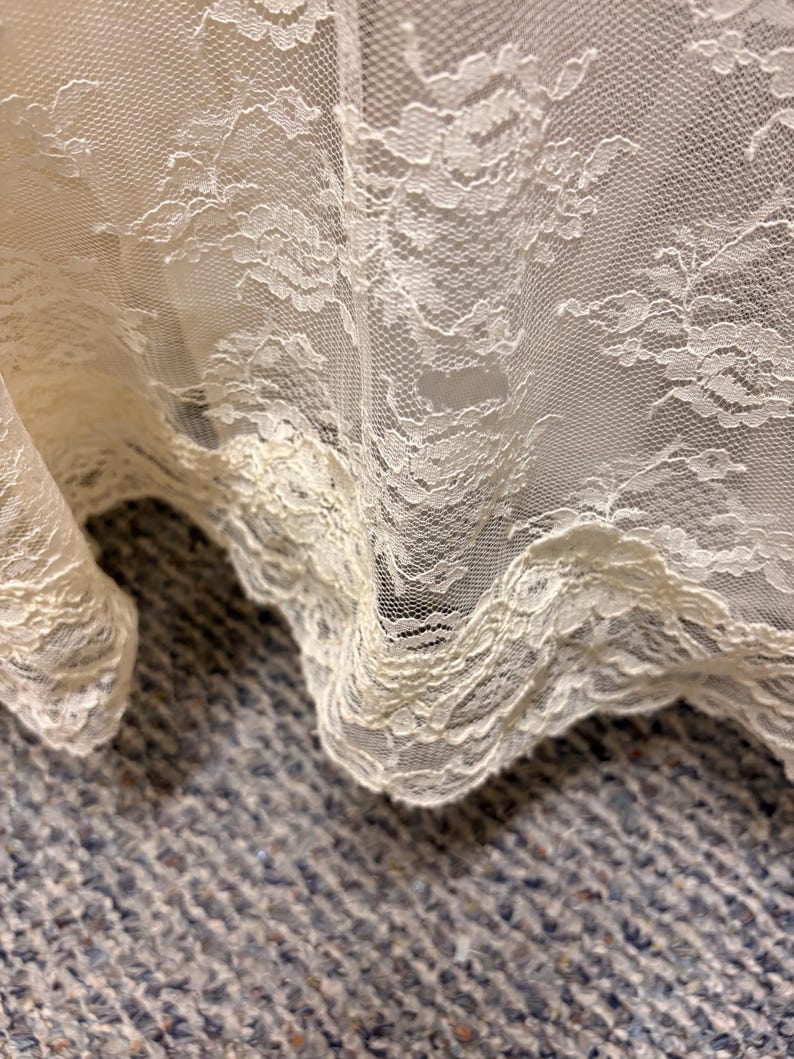 May include: Close-up of ivory lace fabric with a floral pattern. The delicate lace is draped, showing the intricate details and texture. The edges are scalloped, adding a decorative touch. The fabric is likely used for apparel or home decor.
