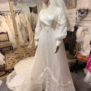 May include: A vintage white wedding dress on a mannequin. The dress features a high neckline, long sleeves, and intricate lace detailing. A long veil and train complete the bridal look. The dress is displayed in a bridal shop setting.