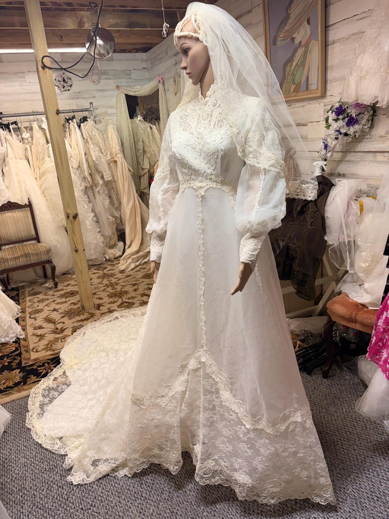 May include: A vintage white wedding dress on a mannequin. The dress features a high neckline, long sleeves, and intricate lace detailing. A long veil and train complete the bridal look. The dress is displayed in a bridal shop setting.