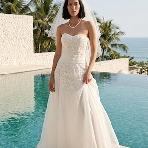May include: A strapless white wedding dress featuring a sweetheart neckline, a fitted bodice with intricate floral lace embroidery, a wide waistband, and a flowing floor-length skirt. A sheer white veil with a pearl-adorned headband is worn.