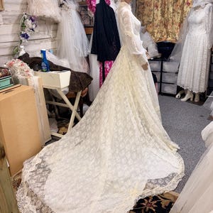 May include: A vintage ivory lace wedding dress on a mannequin. The dress features long sleeves, a high neckline, and a long train. The lace has a floral pattern, and the dress is displayed in a bridal shop setting.