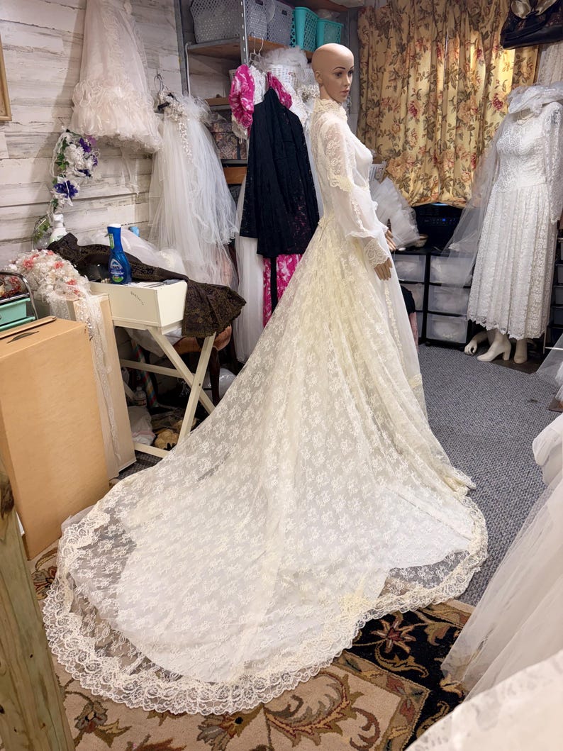 May include: A vintage ivory lace wedding dress on a mannequin. The dress features long sleeves, a high neckline, and a long train. The lace has a floral pattern, and the dress is displayed in a bridal shop setting.