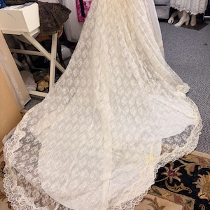 May include: Ivory lace wedding gown with a long train. The dress features a delicate floral pattern and a scalloped lace trim along the hemline. The gown is displayed on a mannequin, showcasing its flowing silhouette and intricate details.