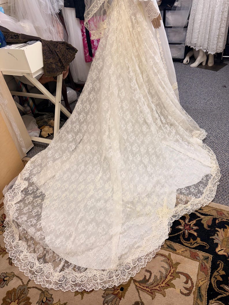 May include: Ivory lace wedding gown with a long train. The dress features a delicate floral pattern and a scalloped lace trim along the hemline. The gown is displayed on a mannequin, showcasing its flowing silhouette and intricate details.