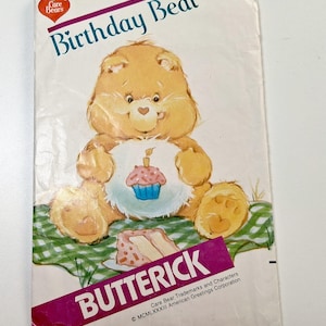 May include: A vintage Butterick sewing pattern envelope for a Care Bears Birthday Bear. The front features a yellow bear with a cupcake graphic on its belly, sitting on a green and white checkered cloth with a slice of cake, labeled 6229.