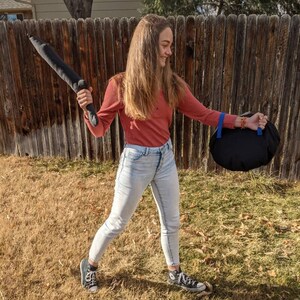 May include: A long-sleeved rust-colored shirt, light-wash denim jeans, and black canvas sneakers. The outfit includes a dark gray plush sword-shaped toy and a round black bag with blue straps, worn while standing outdoors in a grassy area.