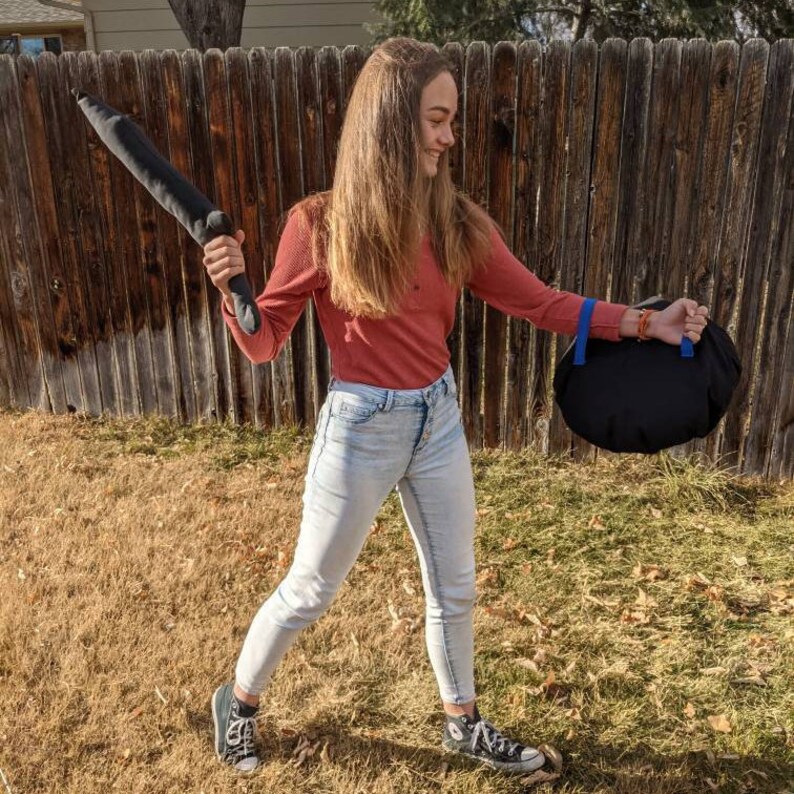 May include: A long-sleeved rust-colored shirt, light-wash denim jeans, and black canvas sneakers. The outfit includes a dark gray plush sword-shaped toy and a round black bag with blue straps, worn while standing outdoors in a grassy area.
