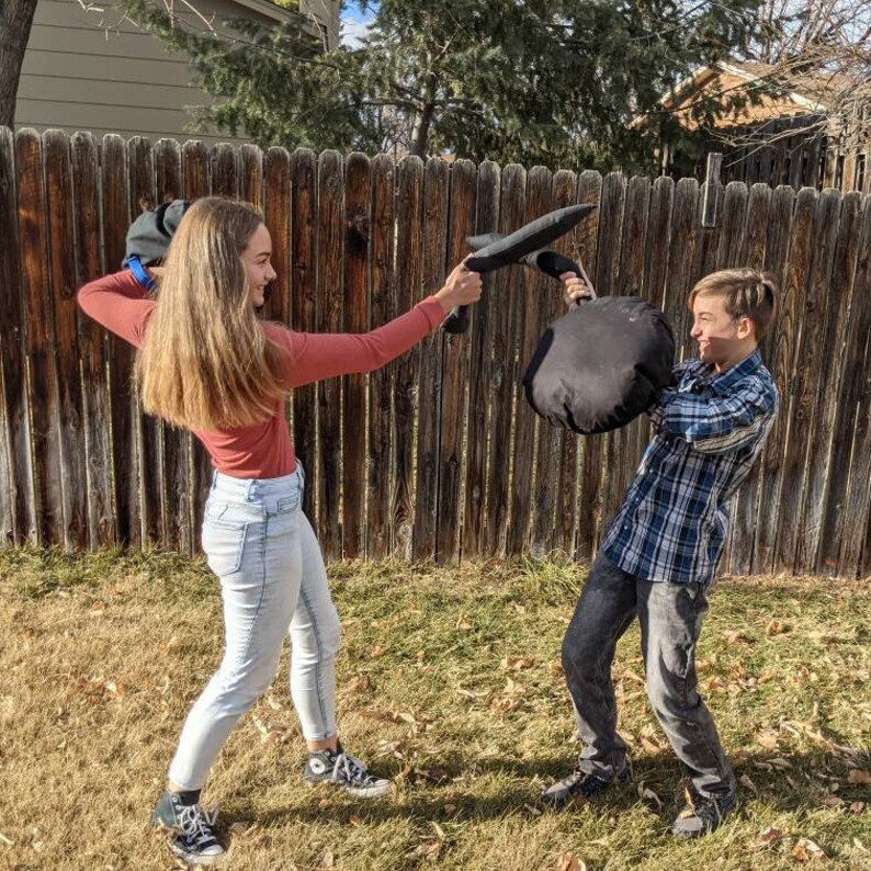 May include: Two individuals engaged in a playful mock sword fight using soft, black fabric-covered foam props. One person wears a long-sleeved rust-colored shirt and light blue jeans, while the other wears a blue plaid shirt and dark gray jeans.