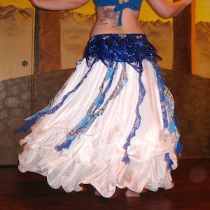 Puede incluir: Una falda blanca de capas con detalles azules y plateados. La falda está hecha de una tela fluida y tiene un dobladillo con volantes. La falda se lleva sobre un cinturón azul y plateado con cuentas.