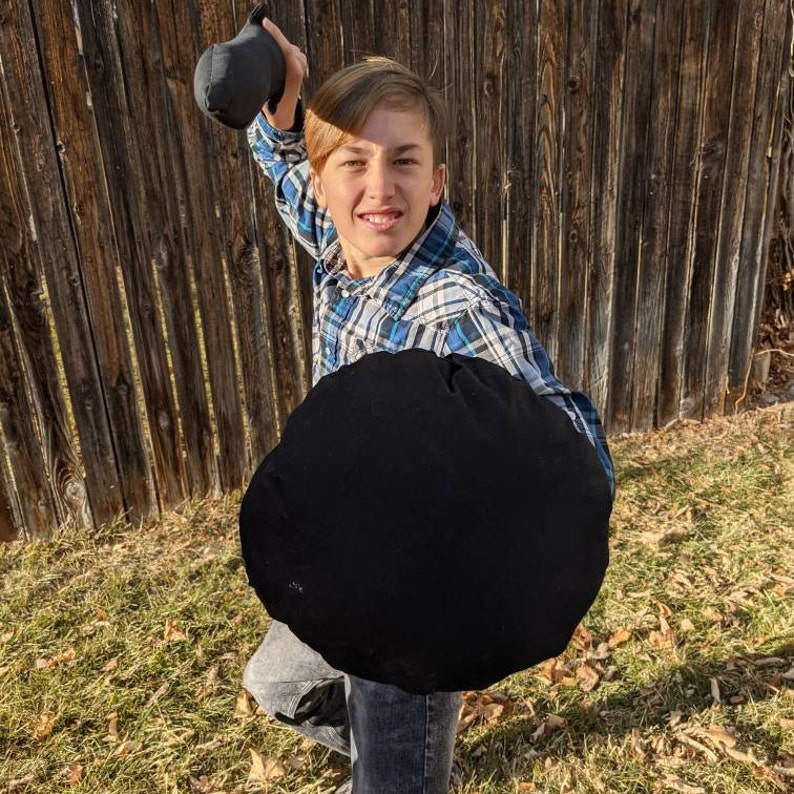 May include: A person wearing a blue and white plaid long-sleeve button-down shirt and gray jeans, holding a solid black circular shield and a black sword-shaped object, both made of a soft fabric material, in a dynamic pose outdoors.