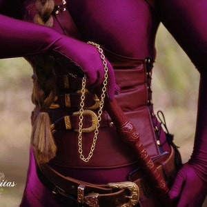 May include: A woman wearing a purple outfit with a gold chain and a brown leather belt. She is holding a brown leather whip.
