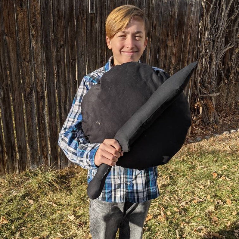 May include: A black fabric shield and a matching black fabric sword. The shield is circular in shape, and the sword features a long, pointed blade with a handle. Both items are constructed from soft, solid black material with visible seams.