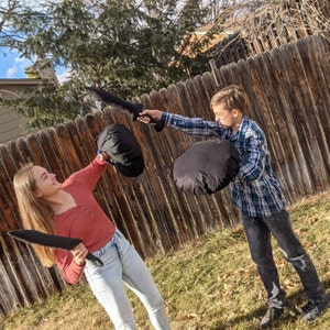 May include: Two black foam swords and two black circular foam shields are being used in a playful mock battle. The swords are long and tapered, while the shields are round and padded. These soft toy weapons are designed for safe, active play.