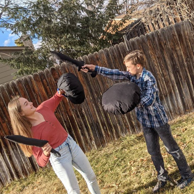 May include: Two black foam swords and two black circular foam shields are being used in a playful mock battle. The swords are long and tapered, while the shields are round and padded. These soft toy weapons are designed for safe, active play.