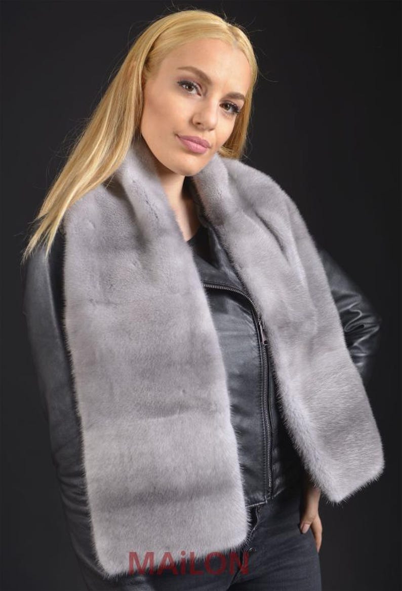 Puede incluir: Una mujer lleva una chaqueta de cuero negra con una bufanda de piel gris claro. La bufanda es larga y se envuelve alrededor del cuello.