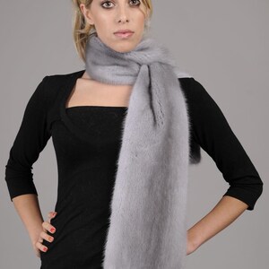 May include: A light gray fur scarf with a long, flowing design. The scarf is worn around the neck of a person wearing a black top and blue jeans.