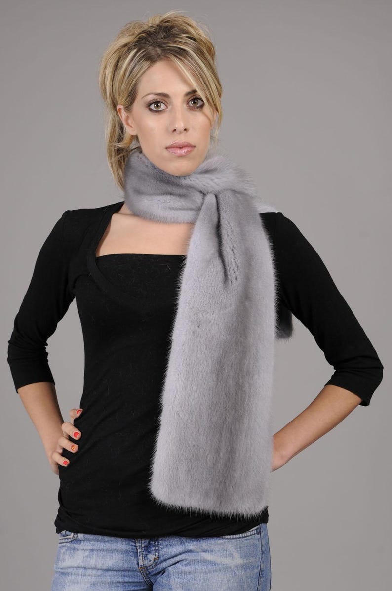 May include: A light gray fur scarf with a long, flowing design. The scarf is worn around the neck of a person wearing a black top and blue jeans.