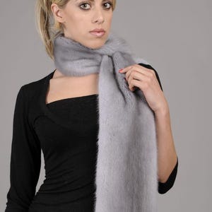 May include: A light gray fur scarf with a long, flowing design. The scarf is worn around the neck of a person with blonde hair.