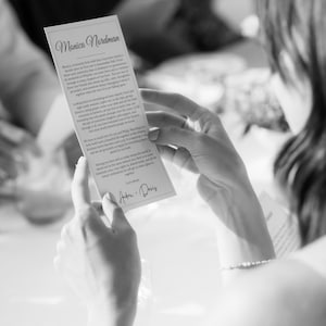 Op de afbeelding: Een zwart-witfoto van een persoon die een rechthoekige kaart met tekst vasthoudt. De kaart is getiteld "Monica Nordman" en bevat een handgeschreven handtekening. De persoon zit aan een tafel, de achtergrond is wazig.