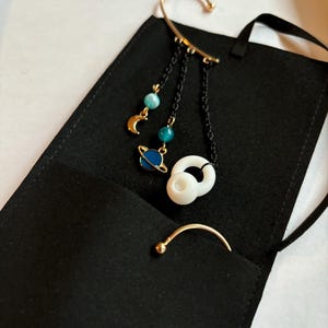 May include: A gold ear cuff with a dangling chain featuring celestial charms, including a crescent moon, a planet with rings, and blue and green beads. The ear cuff is displayed on a black velvet pouch.