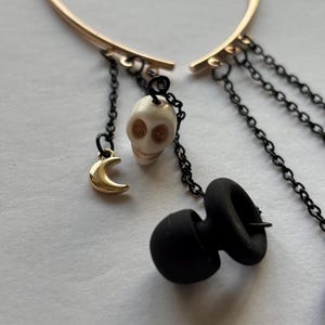 Puede incluir: Puño de oreja en forma de corazón dorado con detalles de cadena negra. Las cadenas cuelgan pequeños amuletos, incluyendo una calavera, una luna creciente y una forma negra redondeada. El puño de oreja está diseñado para ser usado en la oreja.