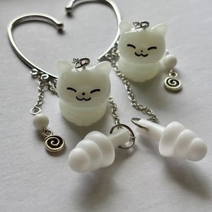 May include: A pair of silver-toned ear cuffs featuring dangling white cat charms with black facial features. Each cuff includes a silver chain, a small white bead, a silver spiral charm, and a white, tiered, conical earplug attachment.