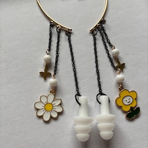 Puede incluir: Un par de pendientes de aro de oro con cadenas colgantes y abalorios. Los abalorios incluyen cuentas blancas, pequeñas mariposas doradas y flores blancas y amarillas. Los pendientes también vienen con un par de tapones para los oídos de silicona blanca.