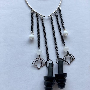 Puede incluir: Pendientes de puño con una parte superior plateada en forma de corazón. Cadenas negras cuelgan con cuentas blancas y dijes de flor de loto. Tapones para los oídos negros cuelgan debajo. Un accesorio único.