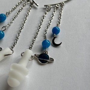May include: A silver-toned ear cuff featuring multiple dangling chains. The chains are adorned with small blue beads, a silver crescent moon charm, a dark blue planet charm with rings, and white, smooth, mushroom-shaped charms.