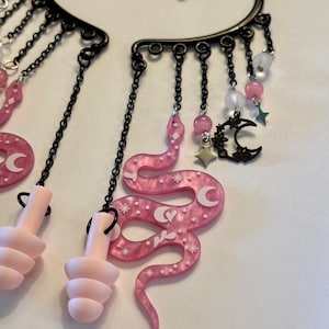 May include: A pair of black metal ear cuffs featuring dangling chains with pink marbled resin snake charms, light pink earplugs, small pink and white beads, and black metal crescent moon and star charms with white celestial printed details.
