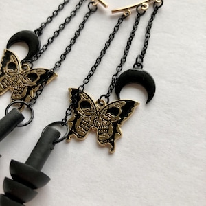 Puede incluir: Pendiente de aro dorado con cadenas y dijes negros. Los dijes incluyen una mariposa de calavera, una luna creciente y un par de tapones para los oídos negros.