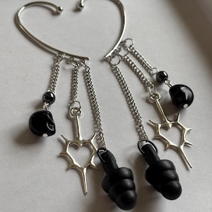 May include: A pair of silver ear cuffs with dangling charms. The charms include black skulls, a heart-shaped charm with spikes, and black earplugs.
