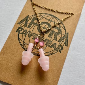 Puede incluir: Un collar de cadena dorada con tapones para los oídos rosas colgando. Los tapones para los oídos son de color rosa claro y tienen un diseño acanalado. El collar está sobre una tarjeta marrón con el texto "AFORA RAVEWEAR" impreso.
