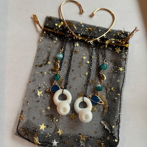 Puede incluir: Un par de pendientes de aro de oro con colgantes, incluyendo planetas, lunas y estrellas. Los colgantes están hechos de esmalte blanco y azul y están unidos a cadenas negras. Los pendientes están expuestos en una bolsa de malla negra y dorada.